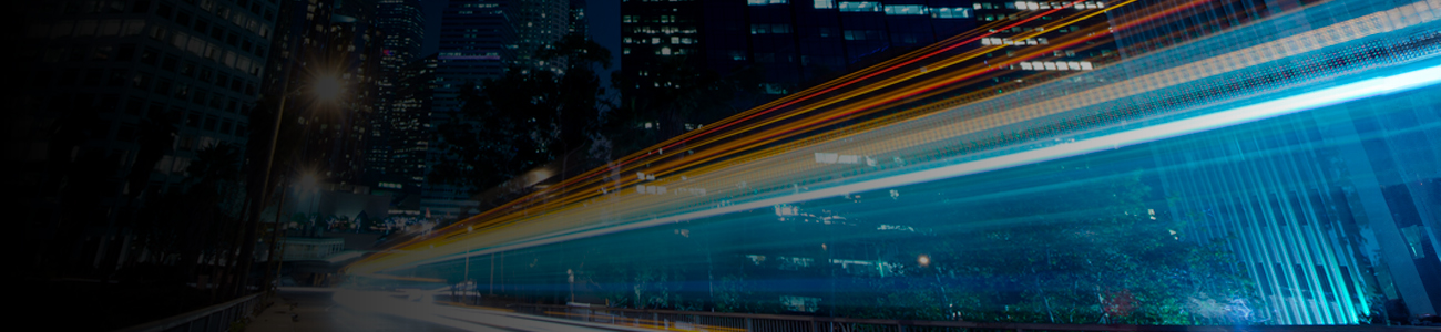 A cityscape at night with skyscrapers in the background. Bright streaks of light from moving vehicles create a dynamic effect in the foreground.