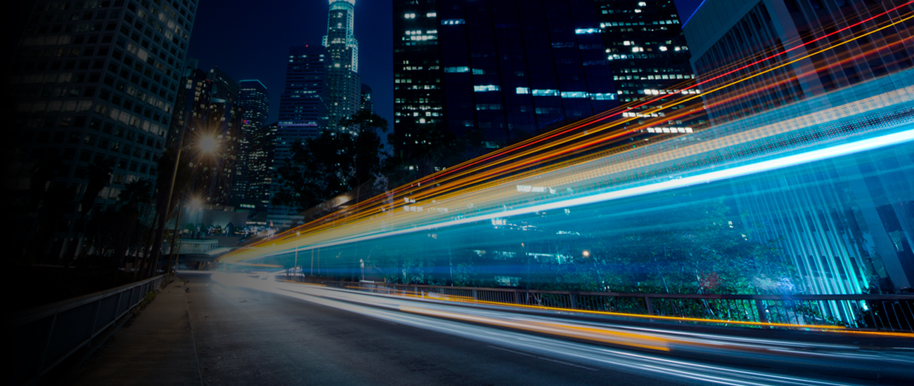 A cityscape at night with skyscrapers in the background. Bright streaks of light from moving vehicles create a dynamic effect in the foreground.